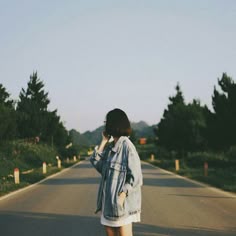 Woman standing on a road surrounded by trees, wearing a denim jacket, holding a phone, suggesting a moment of reflection or communication.