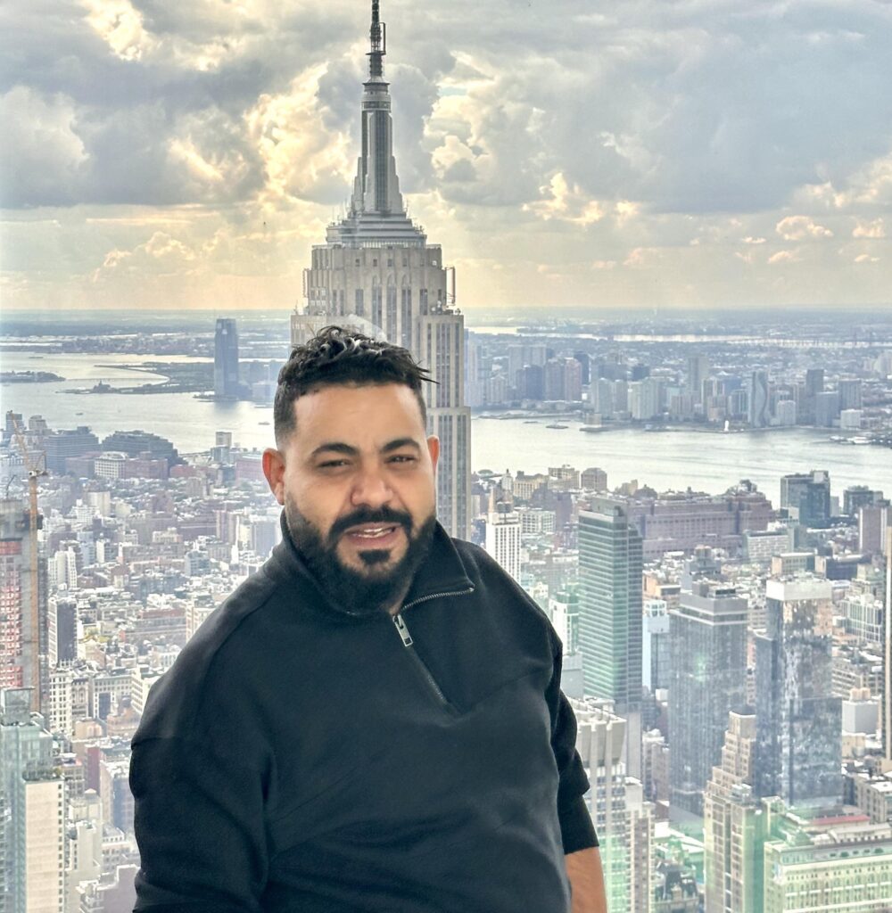 Man in black jacket with a beard standing in front of a city skyline featuring the Empire State Building, emphasizing urban landscapes and personal experiences.