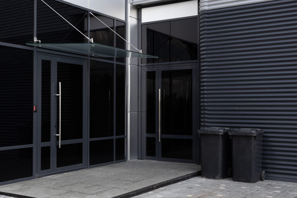 Black glass doors with sleek handles at the entrance of a modern building, featuring a glass awning above and a textured gray wall, highlighting Dr. Glass and Doors' expertise in stylish and secure commercial glass installations.
