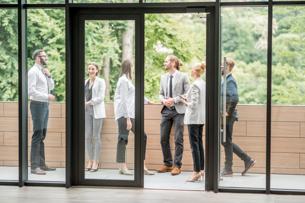 Business professionals engaging in conversation on a modern office balcony, showcasing a collaborative workspace environment with large glass doors and green outdoor views, reflecting themes of commercial glass installation and office design.