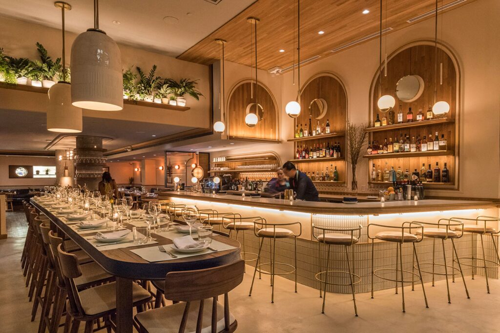 Restaurant interior featuring a long dining table set with glassware and plates, a modern bar with shelves of liquor, decorative lighting, and plants, creating a welcoming atmosphere for dining and socializing.