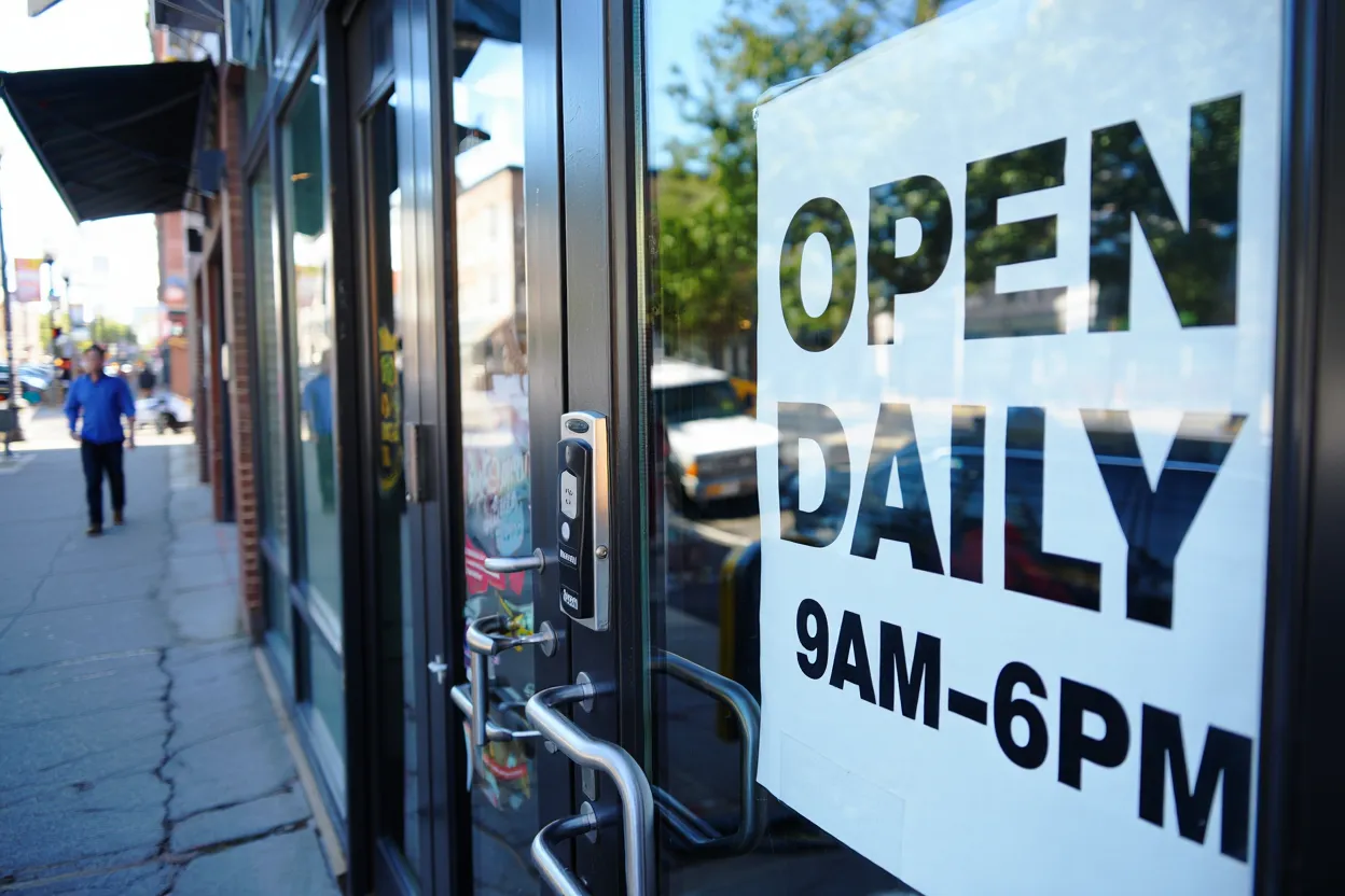 Open sign on commercial glass door indicating daily hours, with a pedestrian walking by on the sidewalk.